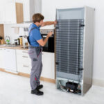 Serviceman In Overall Working On Fridge With Screwdriver In Kitchen