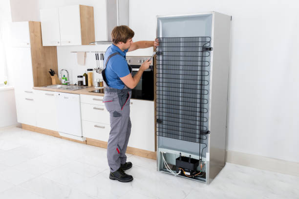 Serviceman In Overall Working On Fridge With Screwdriver In Kitchen Serviceman In Overall Working On Fridge With Screwdriver In Kitchen